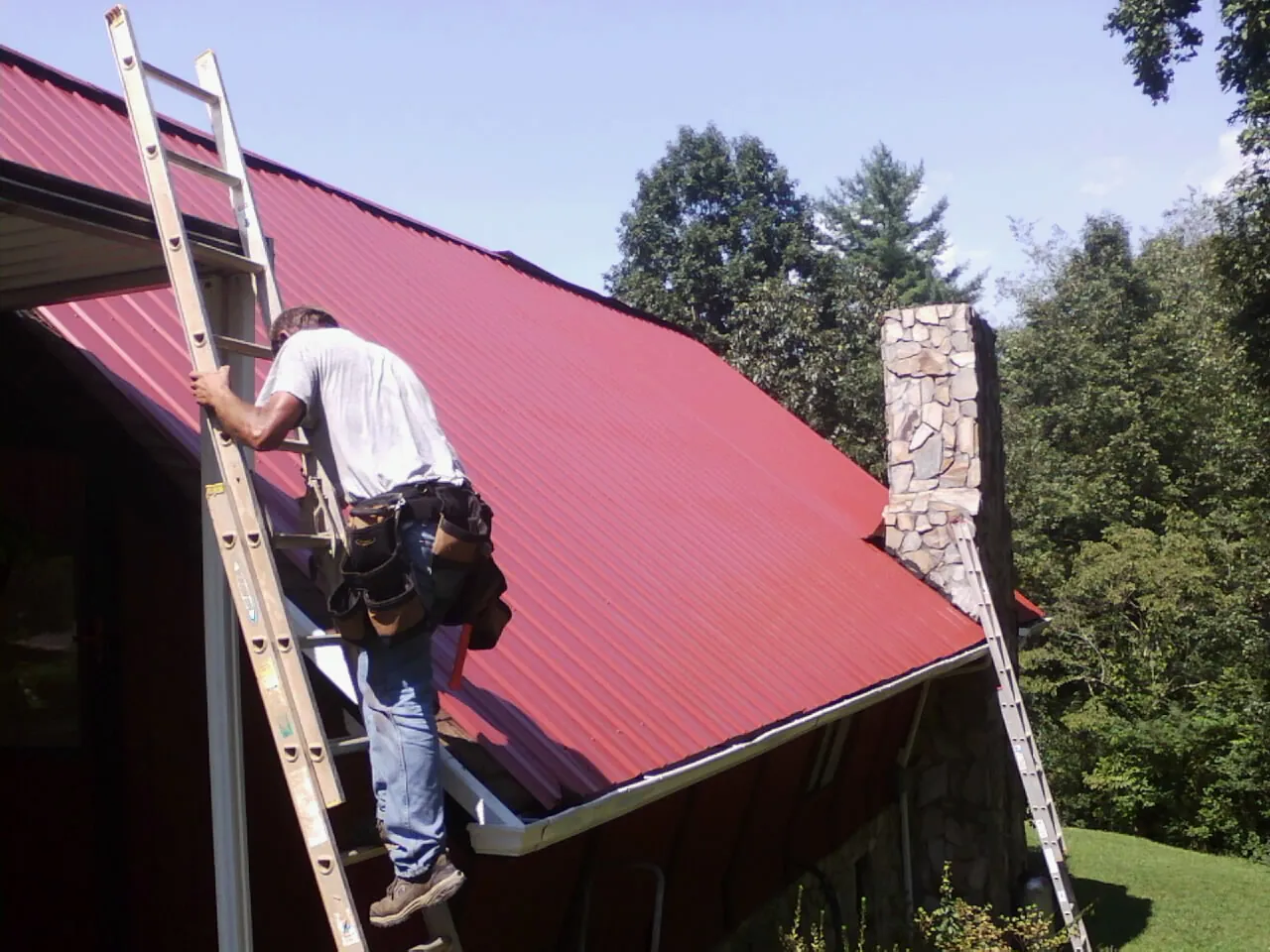 Roofer on a ladder beside a red metal roof and stone chimney — Soffit & Fascia Repair by Roofing Brothers in Oxford